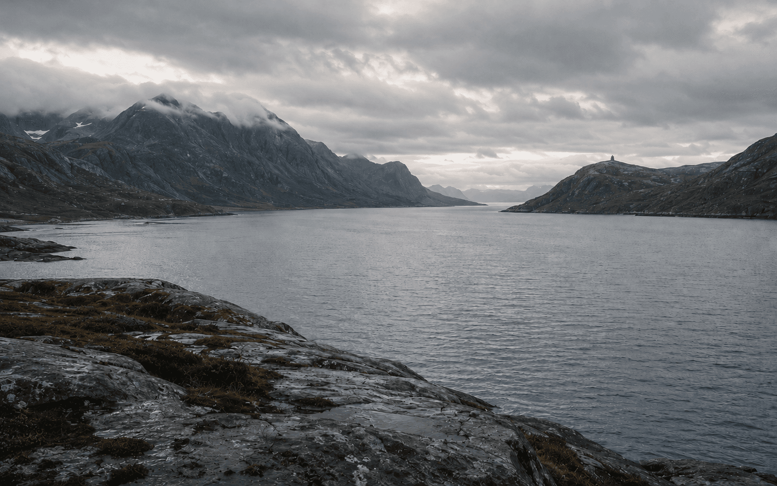 Utsyn over en nordnorsk fjord med fjell og en varde under en overskyet himmel